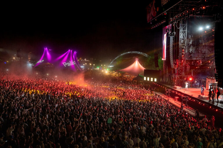 A crowded outdoor music festival at night with a large audience watching a live performance on a stage illuminated by spotlights and adorned with large screens, with event branding visible above the stage.