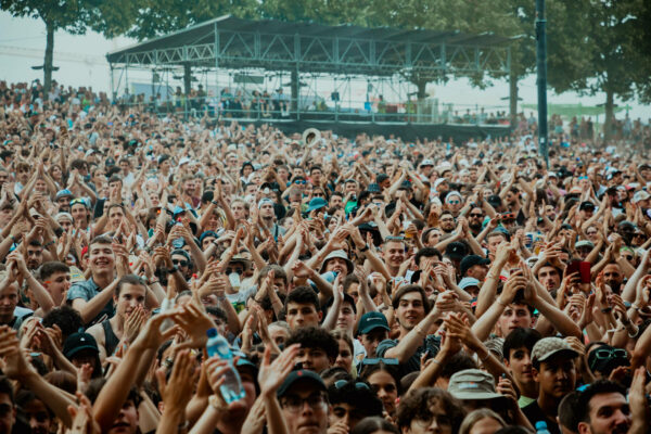 A large crowd of people at an outdoor festival clapping and looking towards a stage, with a few individuals holding up their phones.