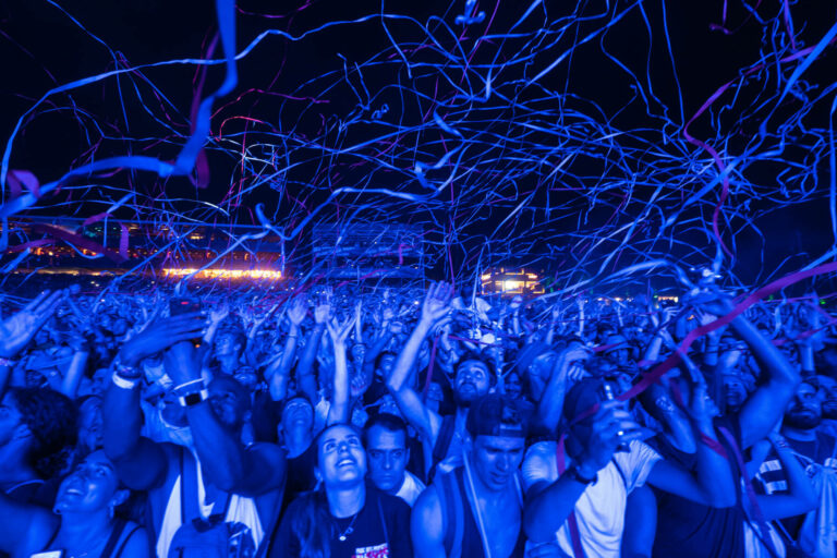 A large crowd at a concert with people cheering and raising their hands, illuminated by blue stage lights with pink streamers flying through the air.