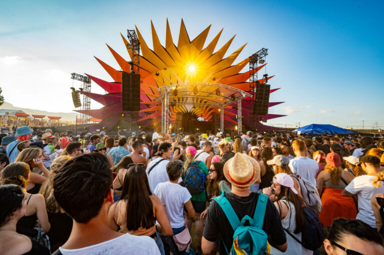 Large crowd of people at an outdoor music festival with a brightly colored stage resembling a sun, bathed in the glow of the setting sun.