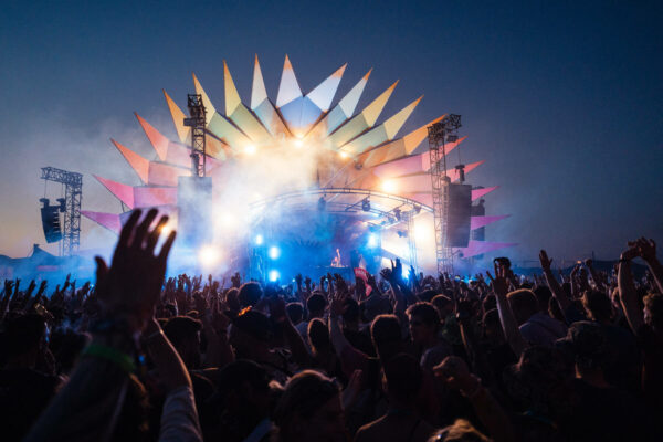 A lively outdoor music festival at dusk with a large crowd of people enjoying themselves, hands raised in the air, in front of a stage with dynamic lighting and a decorative backdrop resembling a sun or flower.