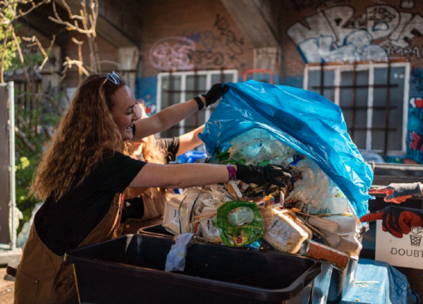 A woman wearing gloves is sorting through a large blue bag of recyclable materials, with a black bin nearby, in a setting illuminated by warm light with graffiti visible in the background.