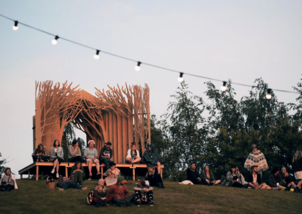 People relaxing on benches and grass near an artistic wooden pavilion under a string of lights at dusk.