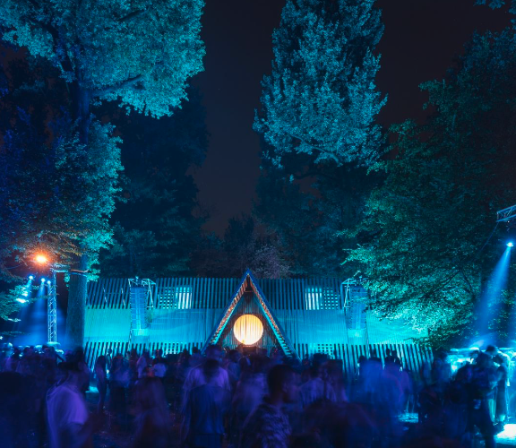 Outdoor night scene at a festival with a crowd of people in motion, illuminated by blue lights, with a triangle-shaped structure and a round light at the center, surrounded by trees.