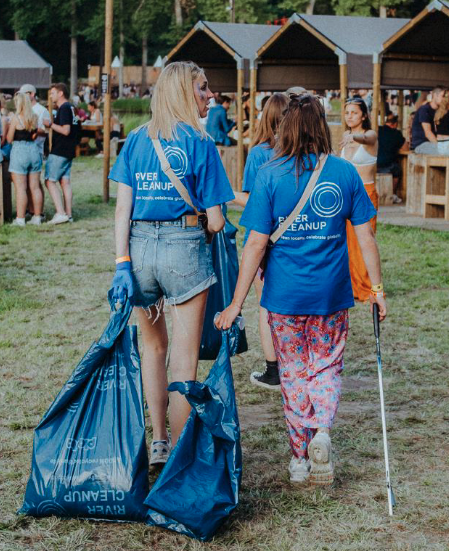 Two volunteers wearing blue River Cleanup t-shirts carry large trash bags and a litter picker at an outdoor event.