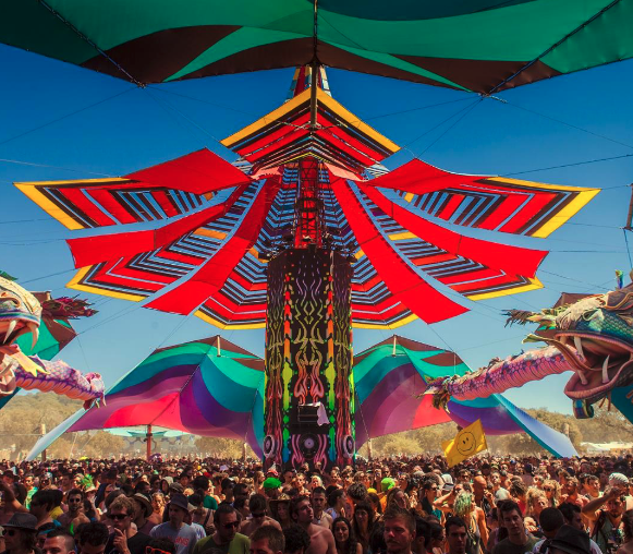 A vibrant outdoor festival scene with a crowd of people under a colorful canopy with a central pillar featuring intricate designs and overhead decorations radiating outwards.