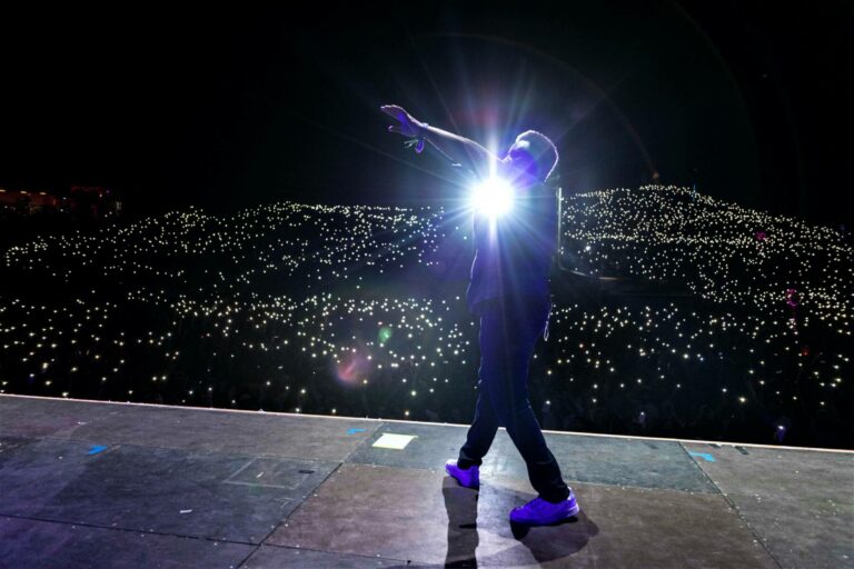 A silhouette of a performer on stage extending their arm towards a bright light, with a sea of twinkling lights from audience members' phones in the background.