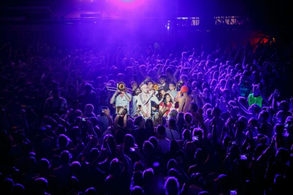 A vibrant concert scene with a large crowd of people surrounding a small group of musicians who are performing among them, illuminated by a spotlight.