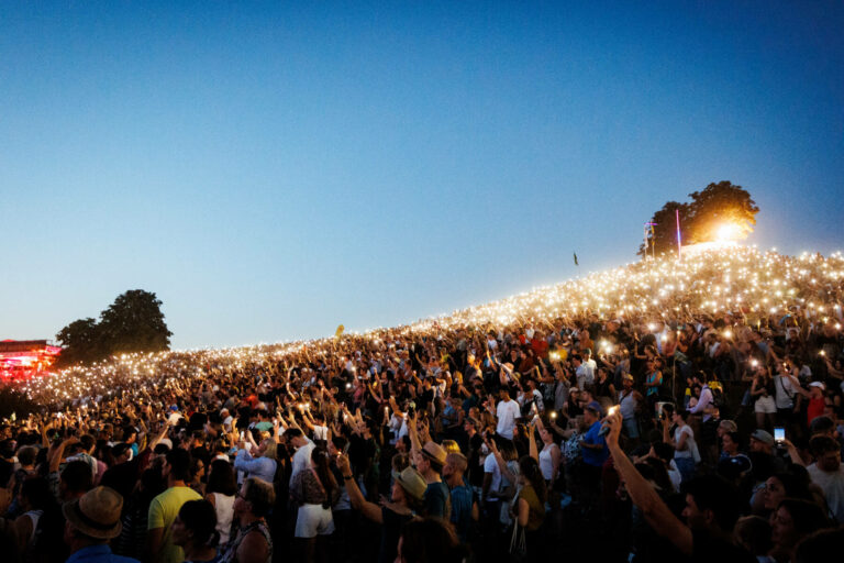 A large crowd of people at an outdoor event, holding up illuminated cell phones in the evening twilight, creating a sea of lights against the darkening sky.