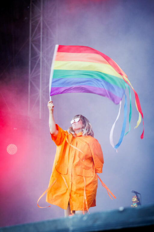A person in an orange raincoat waving a rainbow flag with streamers on a hazy concert stage.