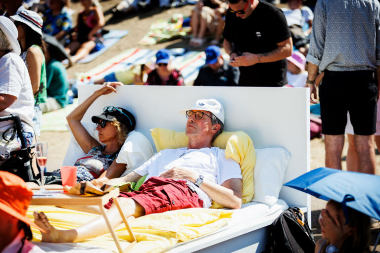A crowded outdoor setting with people lounging and standing around; a man in a white hat and glasses reclines with eyes closed on a makeshift bed in the forefront, next to a seated woman wearing a visor and sunglasses. There's a casual, sunny atmosphere.
