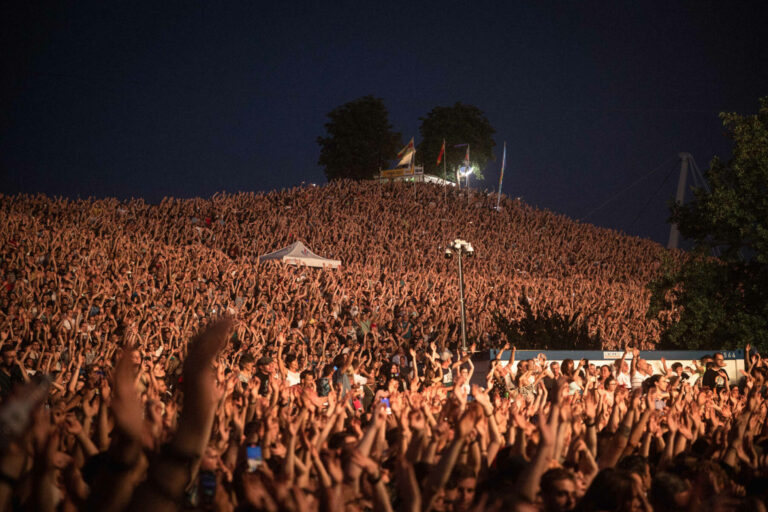 A vast crowd of concertgoers with raised hands, densely packed and cheering at an outdoor music festival during the evening.
