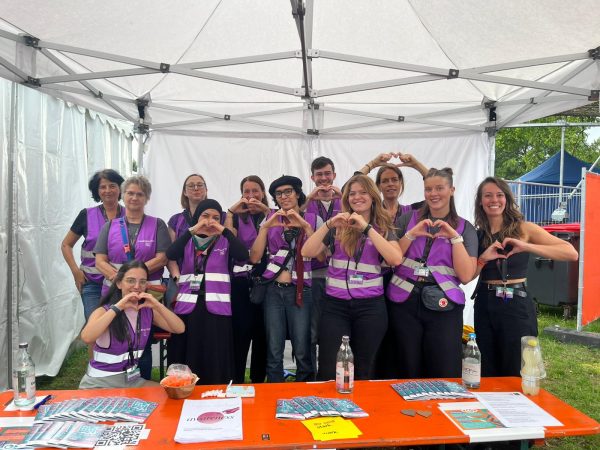 Group of people wearing purple safety vests standing under a white canopy behind a table with pamphlets and bottles, making heart shapes with their hands and smiling at the camera.