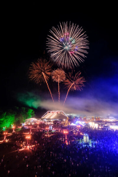 Fireworks display above a crowded festival at night with colorful lights and a large tent in the center.