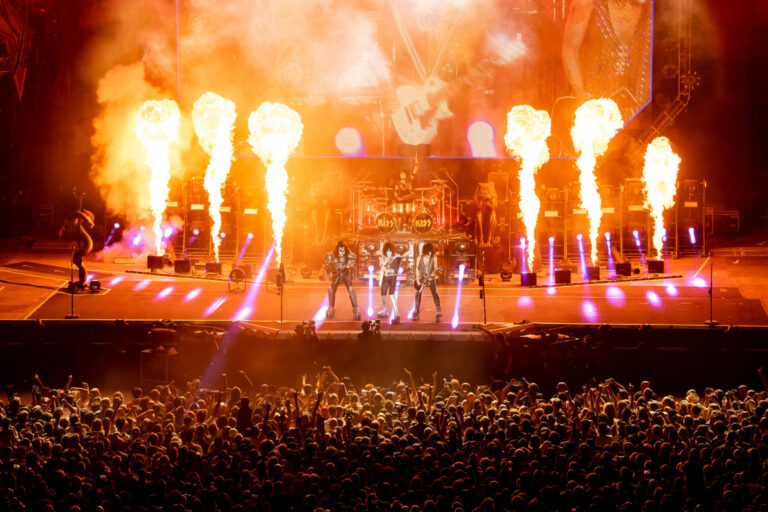 A rock band performing on stage with pyrotechnics, including columns of fire, illuminating them in front of a large, cheering crowd.
