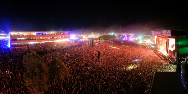 A panoramic view of a crowded outdoor music festival at night featuring a brightly lit stage, multiple screens, and a sea of spectators.