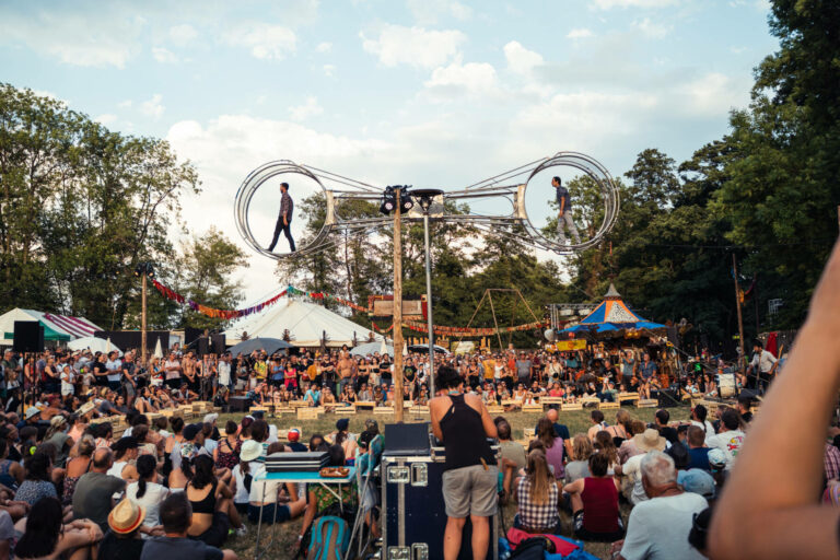 A crowd of spectators watches two performers walking on a large rotating metal wheel installation at an outdoor festival.