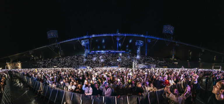A panoramic view of a densely packed outdoor concert at night with a large crowd holding up illuminated mobile phones, creating a sea of lights in front of a large stage structure with lighting rigs.