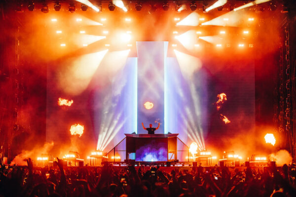 Calvin Harris performing on stage at a concert with hands raised, surrounded by intense stage lighting, plumes of flame, and a crowd of fans.