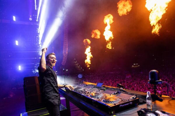 David Guetta raises his fist triumphantly while performing on stage with a crowd in front and flames erupting in the background during a concert at night.