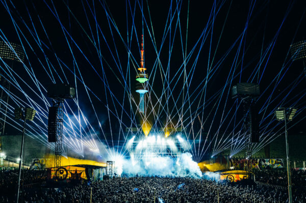 A vibrant outdoor music festival at night with a large crowd, stage lights, and laser beams emanating from the stage towards the sky, with a brightly lit tower in the background.