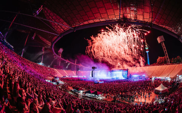 A panoramic view of a large outdoor concert at night with a massive crowd, stage lights, and a vibrant fireworks display above the audience.