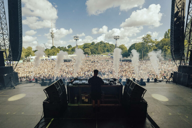 A view from behind a DJ booth on an outdoor festival stage, showing a DJ facing a large crowd with hands raised, and plumes of stage smoke erupting in the air.