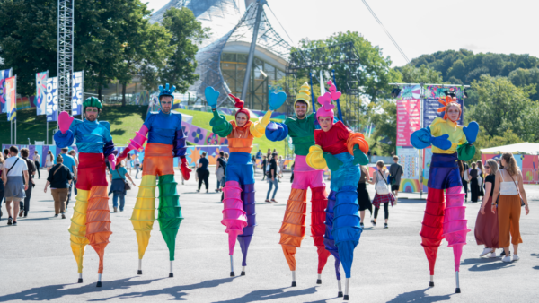 Performers on stilts dressed in colorful, layered costumes resembling twisting, cone-like shapes, with matching oversized gloves and headdresses, participate in a sunny outdoor event.