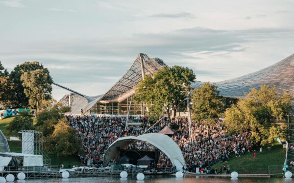 A large outdoor crowd gathered for an event near a modern glass building with a distinctive sloping roof, with a stage set up by the water's edge and trees surrounding the area.