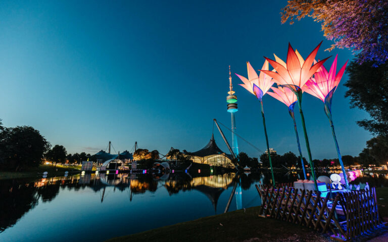 A night scene at a park with illuminated, large, flower-shaped light sculptures by a reflective pond, with a tower and tent-like structures in the background.