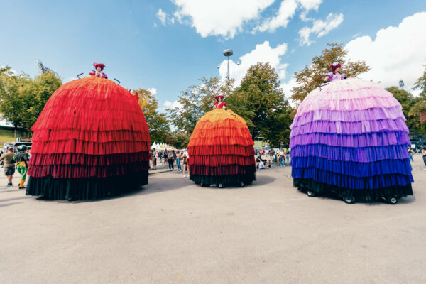 Three large, colorful skirt-shaped costumes on wheels parade through a festival, with performers visible only from the waist up atop the skirts, against a background of a crowd and trees under a blue sky with clouds.