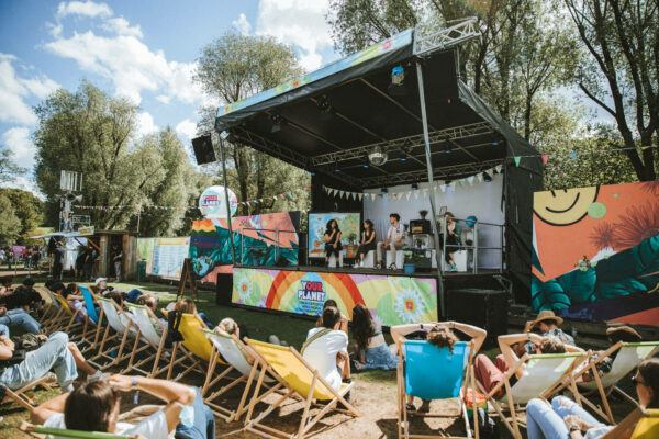 Outdoor festival scene with a small crowd relaxed in deck chairs facing a stage where four people are sitting and talking, surrounded by colorful banners and nature-themed decorations, trees in the background on a sunny day.