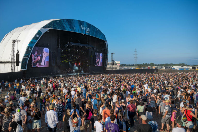 A large crowd of people at an outdoor music festival in front of a stage with a performance displayed on a big screen, under clear blue skies.