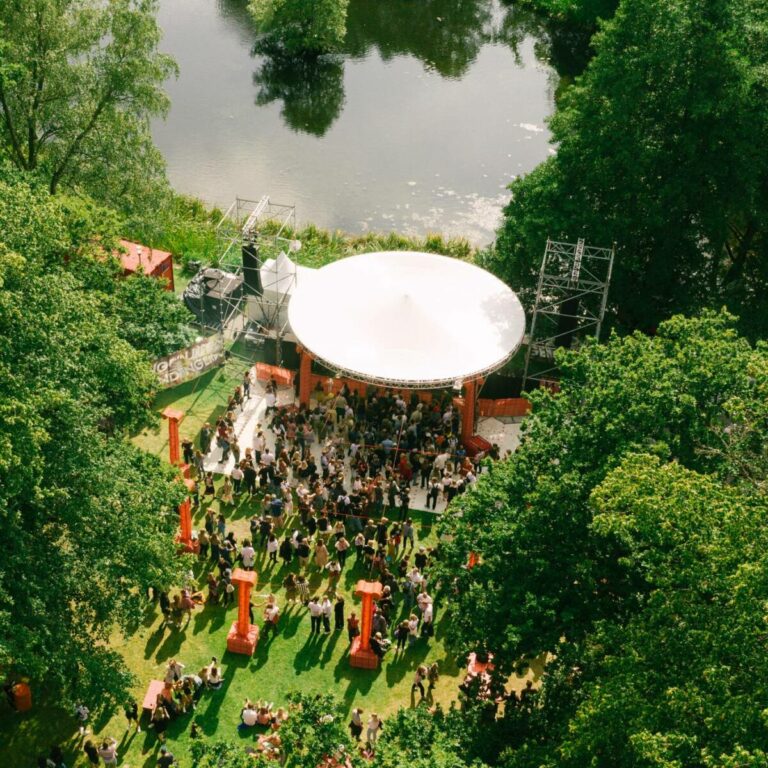 Aerial view of an outdoor event with people gathered under a large white canopy next to a body of water surrounded by lush green trees.