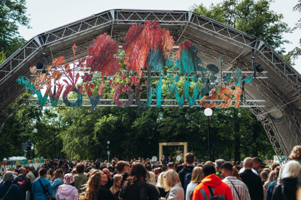 A crowd of people gathered at an outdoor event in front of a stage with colorful, abstract sculptures hanging above.