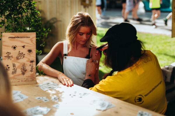 A woman in a white overalls receives a temporary tattoo on her shoulder from an artist wearing a yellow shirt and black cap at an outdoor booth with designs visible on display.