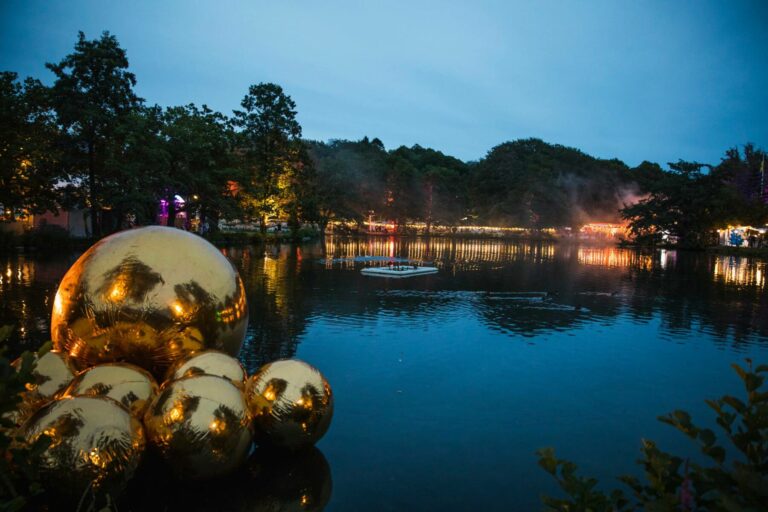 A dusk scene featuring a tranquil lake with a cluster of large, reflective golden spheres in the foreground, and the illuminated banks of the lake lined with trees, lights, and a faint mist in the background. A small floating platform with people is also visible on the water.