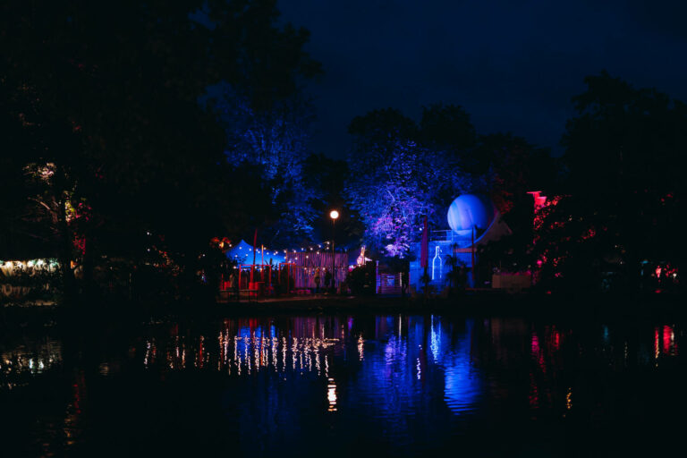 An evening view of a park with colorful lights reflecting on a pond, trees illuminated in blue and red, and a lighted spherical structure in the background.