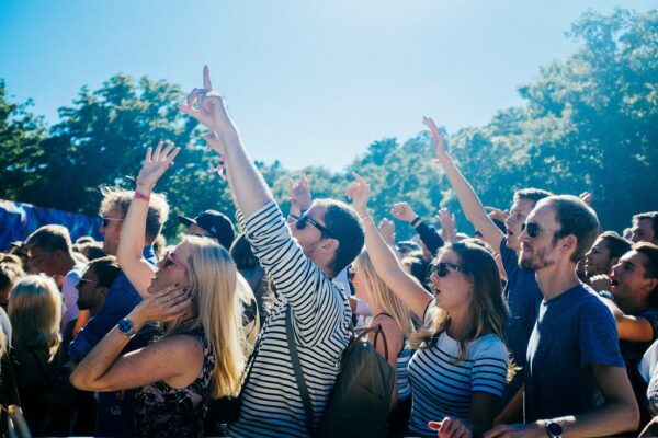 A crowd of cheerful people at an outdoor event, some are raising their hands and pointing upwards, enjoying the sunny weather.