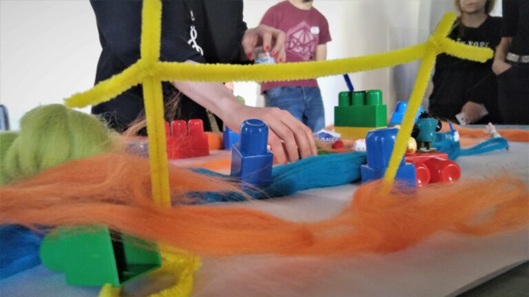 Several people engaging in a hands-on activity with colorful pipes, wool, and construction toys on a table.