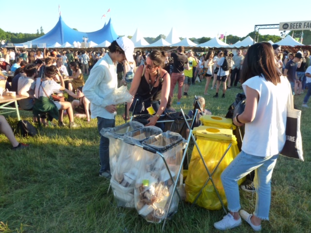 People at an outdoor festival with tents in the background, sorting waste into separate recycling bins under sunlight.