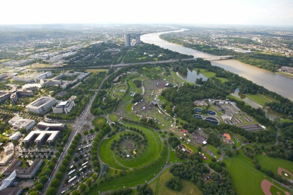 Aerial view of a large park with recreational areas and footpaths, surrounded by buildings, next to a river, with clusters of people gathered for an event.