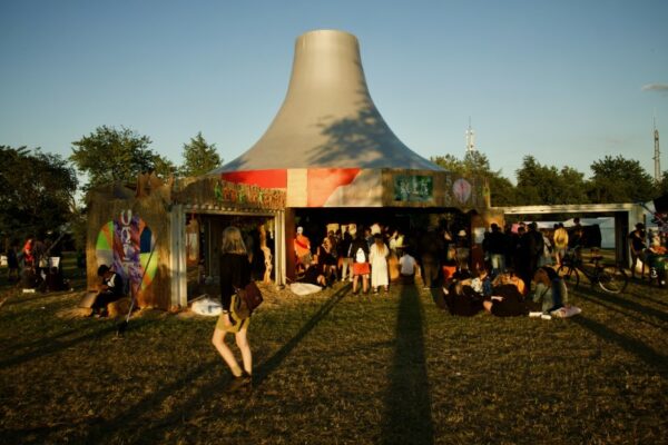 People mingle and relax around a uniquely shaped outdoor structure with colorful decorations at a festival during golden hour.