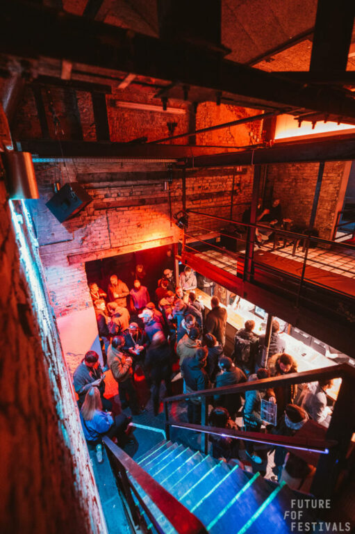 A group of people gathered inside a dimly lit industrial-style building with exposed brick walls and beams, standing on a staircase and a balcony, with a neon sign that reads 
