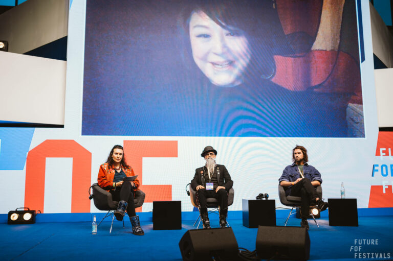 Three individuals are seated on a stage at a panel event, with a large screen behind them displaying a woman's smiling face. The text 