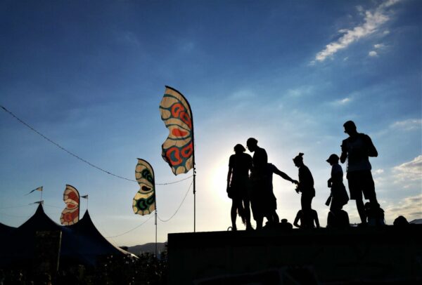 Silhouettes of several people standing and walking on a platform against a bright sky, with colorful flags on poles to the left and a circus tent in the background.