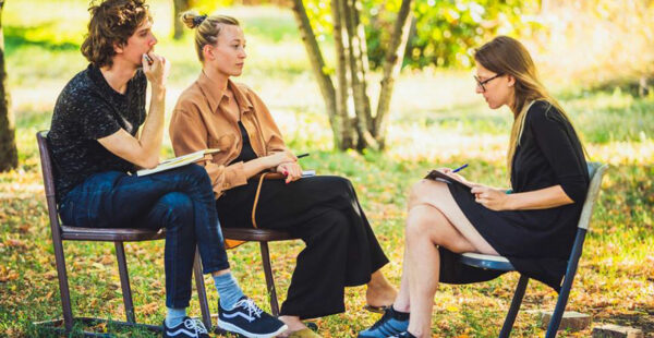 Three people sitting on chairs in an outdoor setting, engaged in a discussion with notepads and pens in hand.