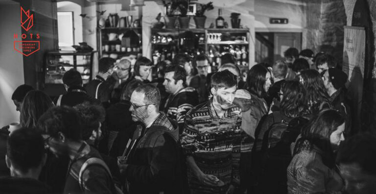 Black and white photo of a crowded indoor event with people socializing, with a logo in the upper left corner reading 