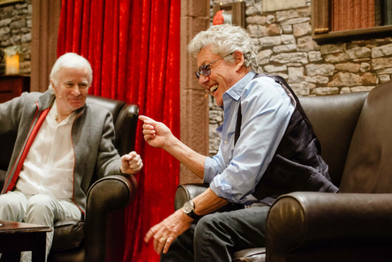 Two elderly men laughing and enjoying a conversation on leather sofas, with one gesturing animatedly and the other sitting back wearing a grey jacket and red scarf. The setting has warm lighting, stone walls, and red curtains in the background.