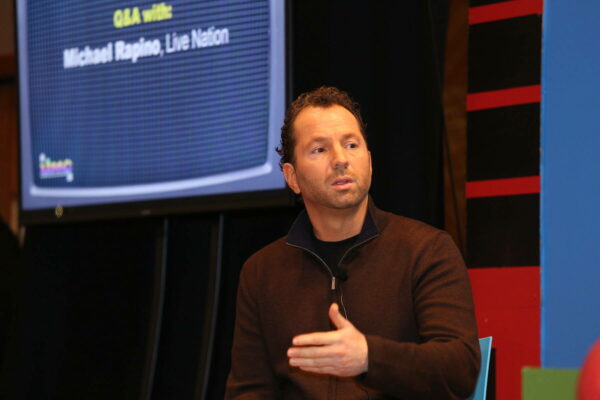 Michael Rapino speaking at a conference with a presentation screen in the background displaying 